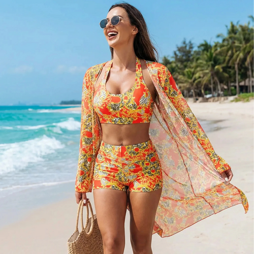 Femme souriante marchant sur une plage de sable fin, portant un ensemble de plage trois pièces aux motifs baroques orange et jaune avec un sac en paille à la main.